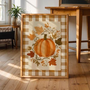 Puede incluir: Una obra de arte enmarcada con temática otoñal, que representa una gran calabaza naranja rodeada de hojas y enredaderas de otoño. La obra está sobre un fondo crema con un borde a cuadros marrón y blanco, todo dentro de un marco de madera.