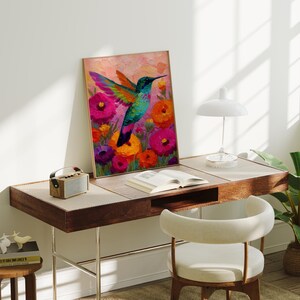 May include: A framed art print depicting a hummingbird with multi-coloured wings, set against a backdrop of bright pink, orange, and purple flowers. The artwork is displayed on a wooden desk with a small speaker, a book, and a white lamp.
