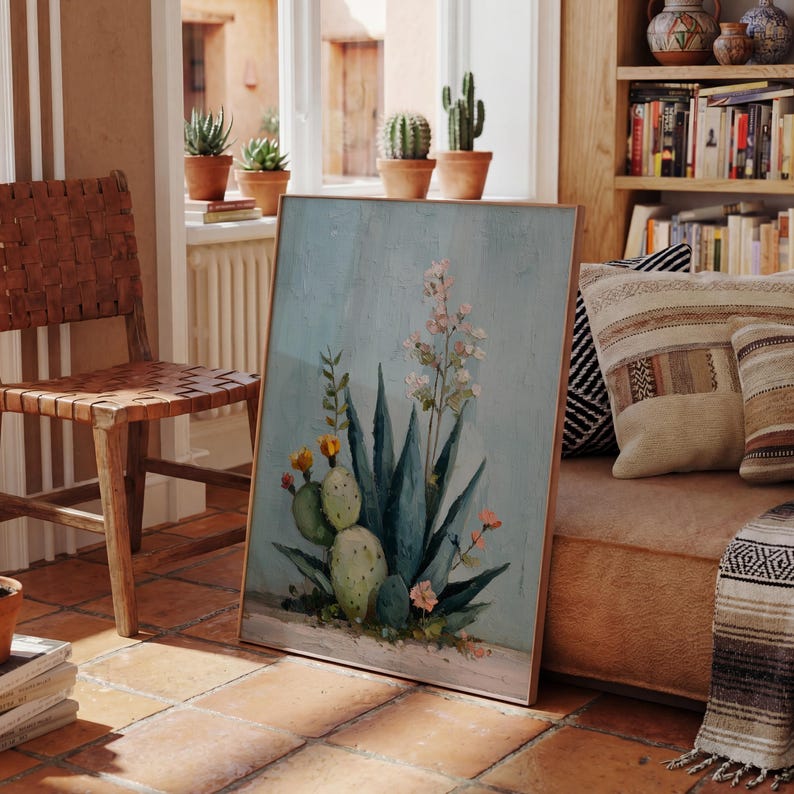 May include: A framed botanical painting featuring a detailed depiction of a desert plant with blue-green leaves, yellow flowers, and pink accents. The artwork is set against a light blue background and has a wooden frame. The scene is in a room with a leather chair and terracotta tile flooring.