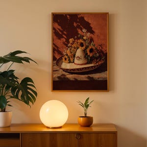 May include: A framed still life painting depicts a sombrero filled with sunflowers and other flowers. The painting hangs above a wooden cabinet. A round, white globe lamp and a small potted plant sit on the cabinet.