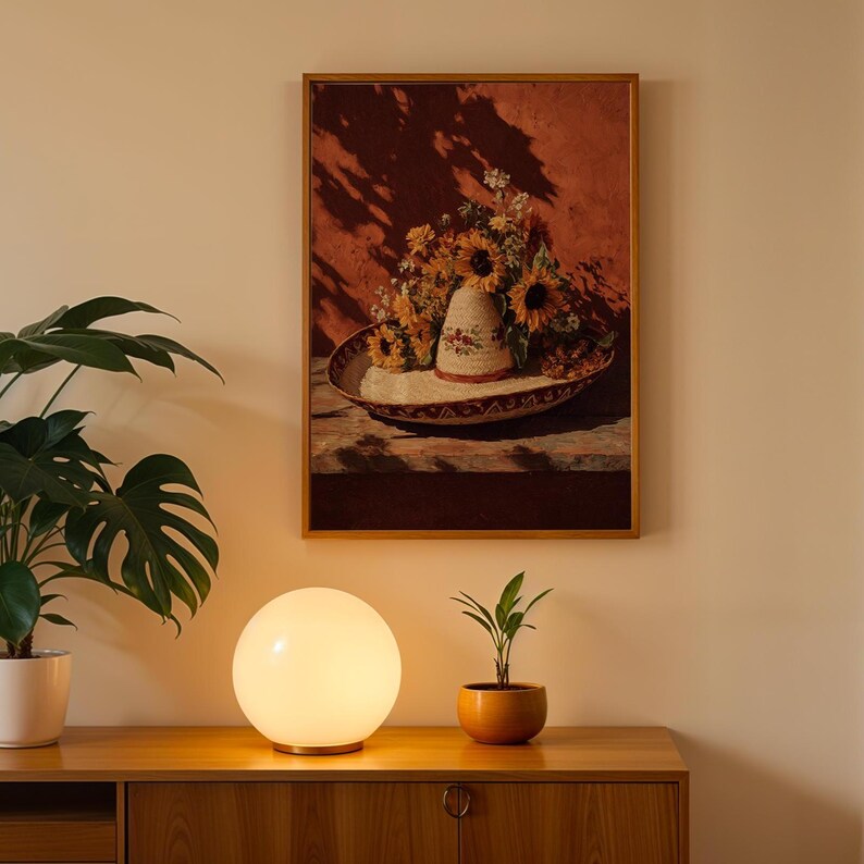 May include: A framed still life painting depicts a sombrero filled with sunflowers and other flowers. The painting hangs above a wooden cabinet. A round, white globe lamp and a small potted plant sit on the cabinet.