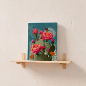 Pode incluir: Impress&atilde;o em tela com um cacto estilizado com grandes flores rosa e borboletas laranja sobre um fundo azul-petr&oacute;leo. A obra de arte &eacute; exibida em uma prateleira de madeira clara, criando um elemento decorativo de parede.