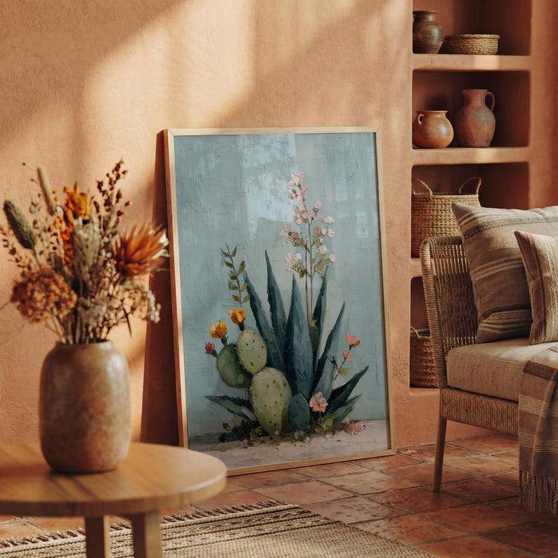 May include: A framed botanical painting featuring a blue agave plant with green cacti and delicate pink and yellow flowers. The artwork is displayed in a warm-toned room with a vase of dried flowers and a woven chair.