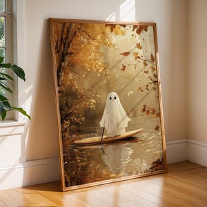 May include: A framed art print depicts a whimsical ghost on a paddleboard in a serene autumn scene. The artwork features a white ghost with black eyes, set against a backdrop of golden trees and a calm body of water. The frame is a warm, natural wood tone.
