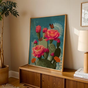 May include: A framed art print featuring a cactus with large pink flowers and orange accents against a teal background. Butterflies are scattered around the cactus. The artwork is in a gold frame and is displayed on a wooden cabinet.