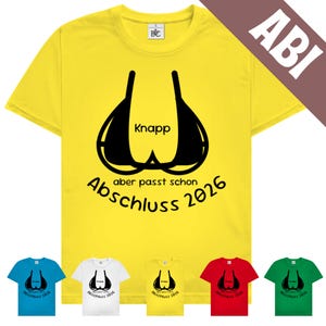 Könnte beinhalten: Gelbes T-Shirt mit schwarzer BH-Grafik und dem Wort "Knapp" darüber. Unter der BH-Grafik steht der Text "Abschluss 2026" und "aber passt schon". Mehrere andere T-Shirts in verschiedenen Farben werden darunter angezeigt.