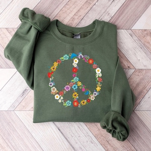 May include: An olive green sweatshirt featuring a colorful floral peace sign design. The peace sign is composed of various flowers in shades of red, yellow, blue, and white. The sweatshirt has long sleeves and a crew neck.