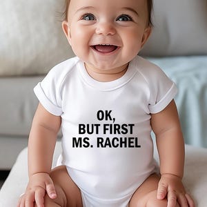 May include: A smiling baby wearing a white onesie with the text "OK, BUT FIRST MS. RACHEL" in black. The baby has brown hair and is sitting on a light-colored surface. The background includes a couch and pillows.