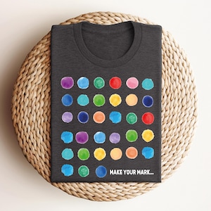 May include: Dark gray t-shirt with a colorful dot pattern. The design features rows of watercolor-style dots in various colors, including blue, green, red, and yellow. The text "MAKE YOUR MARK..." is printed at the bottom of the shirt.