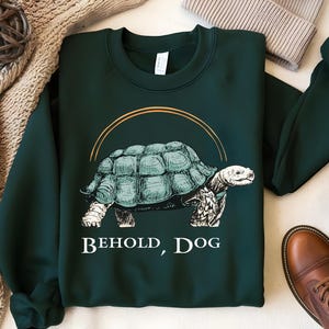 Puede incluir: Una sudadera de color verde oscuro con un gr&aacute;fico de una tortuga con el texto "Behold, Dog" impreso en la parte delantera.