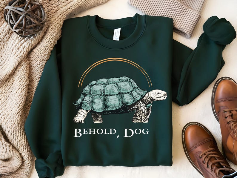 Puede incluir: Una sudadera de color verde oscuro con un gr&aacute;fico de una tortuga con el texto "Behold, Dog" impreso en la parte delantera.