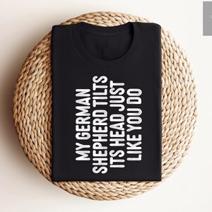 May include: Black t-shirt with white text that reads "My German Shepherd Tilts Its Head Just Like You Do."