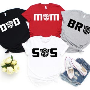 May include: Four t-shirts in black, red, white, and gray with white and black text and graphics. The shirts say "DAD", "MOM", "BR", and "S/S". A pair of white sneakers and denim shorts are also in the image.