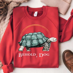 Puede incluir: Sudadera roja con un gr&aacute;fico de una tortuga y el texto "Behold, Dog".