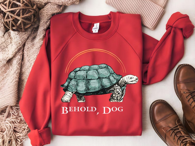 Puede incluir: Sudadera roja con un gr&aacute;fico de una tortuga y el texto "Behold, Dog".