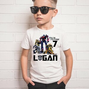 May include: White t-shirt with a Transformers graphic featuring Optimus Prime and other Autobots. The shirt also has the text "Transformers" and the number "7" printed on it. The name "Logan" is printed below the graphic in black.