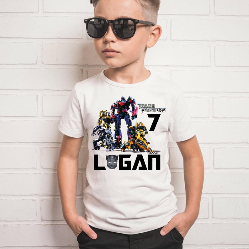 May include: White t-shirt with a Transformers graphic featuring Optimus Prime and other Autobots. The shirt also has the text "Transformers" and the number "7" printed on it. The name "Logan" is printed below the graphic in black.