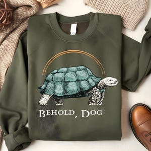 Puede incluir: Una sudadera verde con un dibujo de una tortuga y el texto "Behold, Dog".