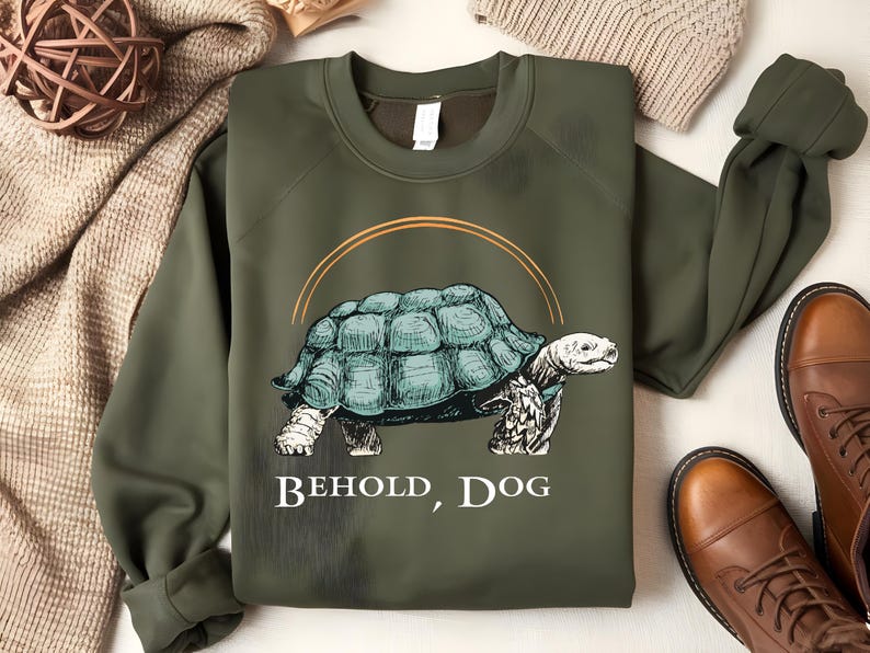 Puede incluir: Una sudadera verde con un dibujo de una tortuga y el texto "Behold, Dog".