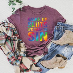 May include: A maroon t-shirt with the text "WAKE UP SLUTS IT'S TIME TO SIN" in rainbow colors. The shirt is paired with blue jeans, tan ankle boots, and a plaid scarf. A sprig of greenery is also present.