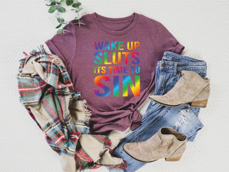 May include: A maroon t-shirt with the text "WAKE UP SLUTS IT'S TIME TO SIN" in rainbow colors. The shirt is paired with blue jeans, tan ankle boots, and a plaid scarf. A sprig of greenery is also present.