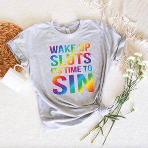 May include: A light gray t-shirt with the text "WAKE UP SLUTS IT'S TIME TO SIN" in rainbow colors. The shirt is knotted at the bottom and is displayed on a white surface with a woven basket and flowers.