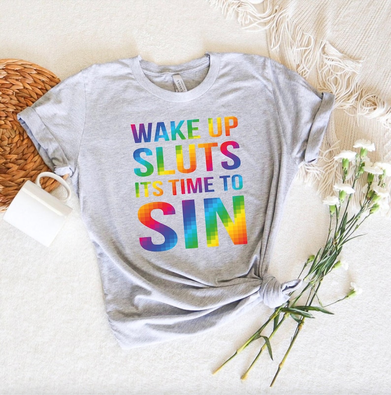 May include: A light gray t-shirt with the text "WAKE UP SLUTS IT'S TIME TO SIN" in rainbow colors. The shirt is knotted at the bottom and is displayed on a white surface with a woven basket and flowers.
