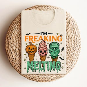 K&ouml;nnte beinhalten: Cremefarbenes T-Shirt mit einem Halloween-Motiv. Das Design zeigt drei Eiswaffeln mit gruseligen Gesichtern: einen K&uuml;rbis, einen Totenkopf und Frankensteins Monster. Der Text "I'M FREAKING MELTING" befindet sich &uuml;ber und unter den Kegeln.