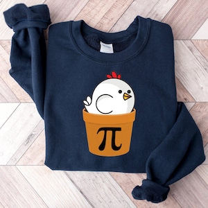 May include: Navy blue sweatshirt featuring a cartoon chicken in a pot with the pi symbol. The chicken is white with a red comb and yellow beak. The pot is orange. A cozy and fun design for casual wear.