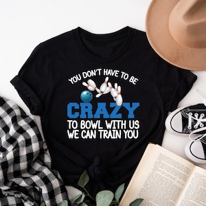 May include: Black t-shirt with white text that reads "You don't have to be crazy to bowl with us, we can train you". The text is surrounded by a graphic of bowling pins and a bowling ball.