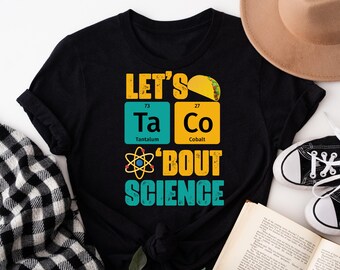May include: Black t-shirt with a graphic design that says "Let's Taco 'Bout Science". The graphic features a taco, the chemical symbols Ta and Co, and an atom.