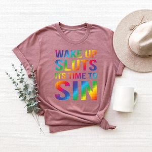 May include: A mauve-colored t-shirt with the text "WAKE UP SLUTS ITS TIME TO SIN" in rainbow-colored block letters. A white mug and a beige hat are also in the image. The shirt is folded at the bottom.
