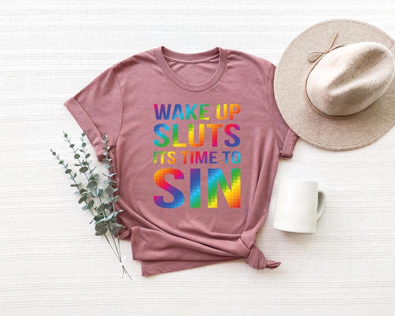 May include: A mauve-colored t-shirt with the text "WAKE UP SLUTS ITS TIME TO SIN" in rainbow-colored block letters. A white mug and a beige hat are also in the image. The shirt is folded at the bottom.
