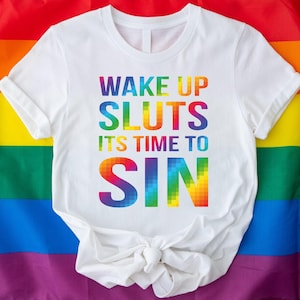 May include: White t-shirt with the text "WAKE UP SLUTS ITS TIME TO SIN" in rainbow colors. The shirt is displayed on a rainbow flag background, representing the LGBTQ+ community.