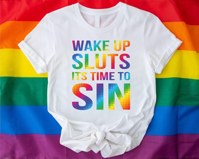 May include: White t-shirt with the text "WAKE UP SLUTS ITS TIME TO SIN" in rainbow colors. The shirt is displayed on a rainbow flag background, representing the LGBTQ+ community.