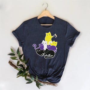 Puede incluir: Camiseta azul marino con un diseño de gatos de dibujos animados amarillos, morados, blancos y negros. Un gato amarillo sostiene una bandera. Los gatos están apilados. La camiseta está colgada de una percha de madera. El diseño está centrado.