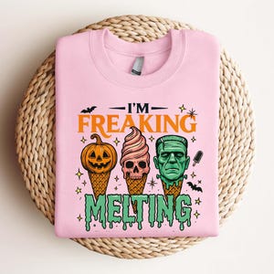 K&ouml;nnte beinhalten: Hellrosa Sweatshirt mit dem Text "I'M FREAKING MELTING". Das Design zeigt einen K&uuml;rbis, einen Totenkopf und Frankensteins Monster als Eist&uuml;ten. Enth&auml;lt Flederm&auml;use, Sterne und tropfenden gr&uuml;nen Text.