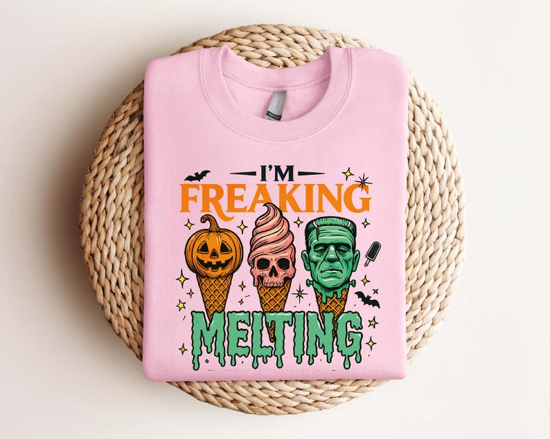 K&ouml;nnte beinhalten: Hellrosa Sweatshirt mit dem Text "I'M FREAKING MELTING". Das Design zeigt einen K&uuml;rbis, einen Totenkopf und Frankensteins Monster als Eist&uuml;ten. Enth&auml;lt Flederm&auml;use, Sterne und tropfenden gr&uuml;nen Text.