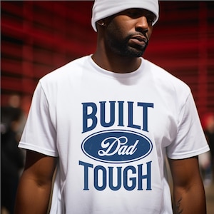 May include: White t-shirt with the words "BUILT Dad TOUGH" in navy blue. The text is arranged in a stacked format, with "Dad" inside an oval shape. The shirt is a crew neck style.