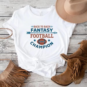 May include: White t-shirt with the text "BACK TO BACK FANTASY FOOTBALL CHAMPION" in blue and red. The design includes a football graphic and an arrow. A brown hat and boots are visible in the background.