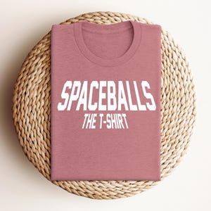 May include: A pink t-shirt with the text "Spaceballs The T-Shirt" in white.