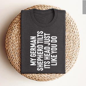 May include: A dark gray t-shirt with white text that reads "My German Shepherd Tilts Its Head Just Like You Do".