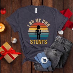 May include: A navy blue t-shirt with a vintage-style graphic of a person with a broken arm and the text "I do my own stunts".