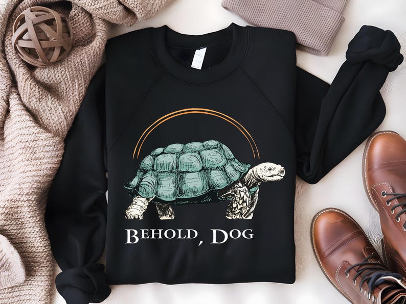 Puede incluir: Sudadera negra con un gr&aacute;fico de una tortuga con el texto "Behold, Dog" impreso en la parte delantera.