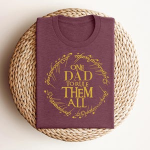 May include: Burgundy t-shirt with a gold inscription that reads "One Dad to Rule Them All" in a circle.