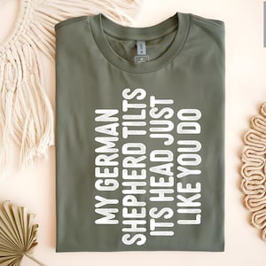 May include: A green t-shirt with white text that reads "My German Shepherd Tilts Its Head Just Like You Do."