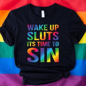 May include: Black t-shirt with the text "WAKE UP SLUTS ITS TIME TO SIN" in rainbow colors. The shirt is displayed on a rainbow flag background. The shirt has short sleeves and a crew neck.