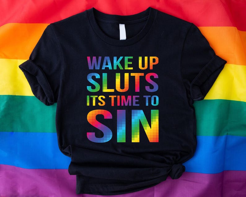 May include: Black t-shirt with the text "WAKE UP SLUTS ITS TIME TO SIN" in rainbow colors. The shirt is displayed on a rainbow flag background. The shirt has short sleeves and a crew neck.