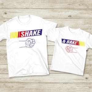 May include: Two white t-shirts with the text "SHAKE" and "& BAKE" in a colorful design. The word "SHAKE" is in blue, red, and yellow, and the word "& BAKE" is in blue and red. Both shirts feature a fist graphic.