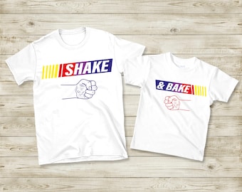 May include: Two white t-shirts with the text "SHAKE" and "& BAKE" in a colorful design. The word "SHAKE" is in blue, red, and yellow, and the word "& BAKE" is in blue and red. Both shirts feature a fist graphic.